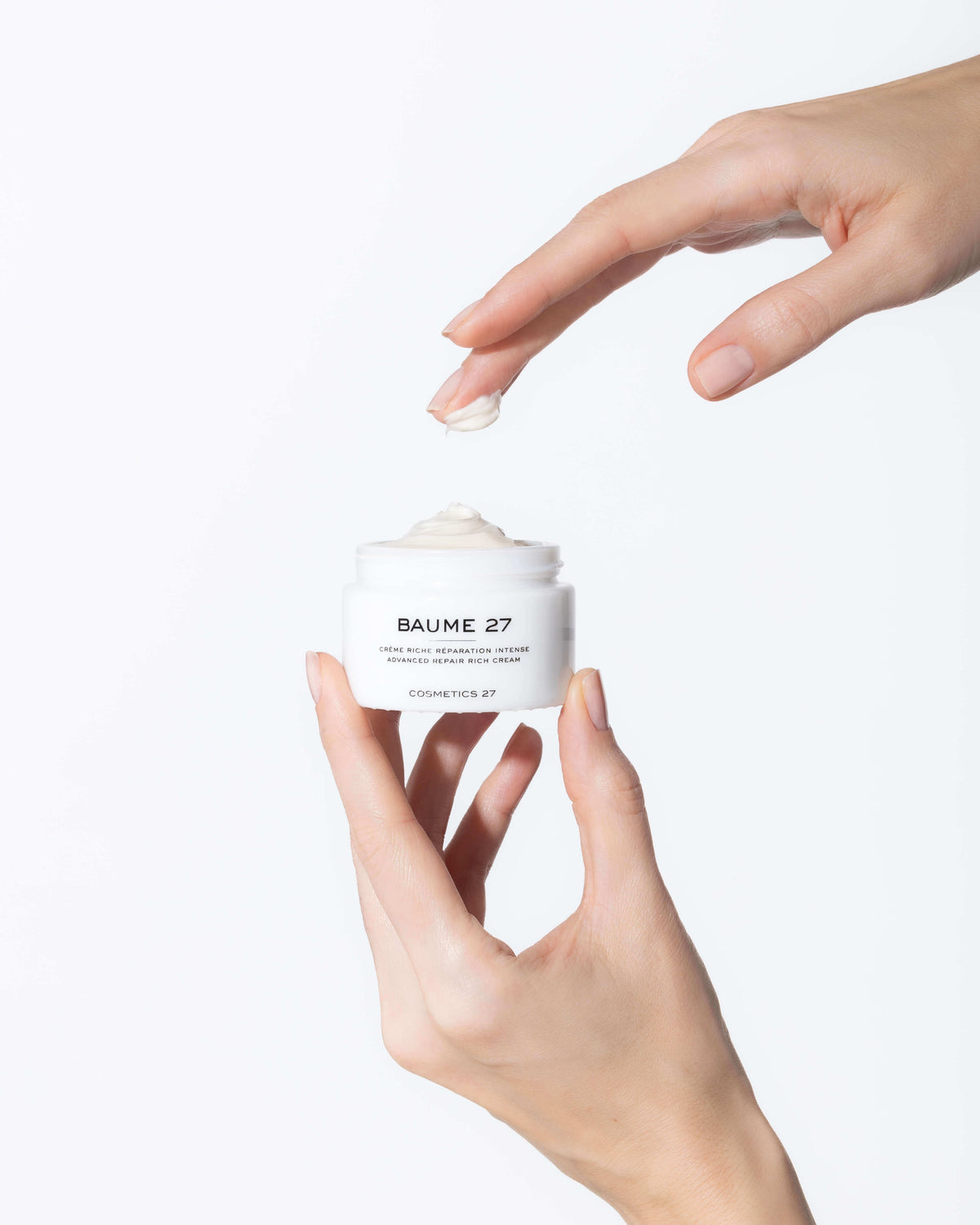 One hand holding an open jar of Baume 27 Rich Cream, the other above it with cream on a fingertip