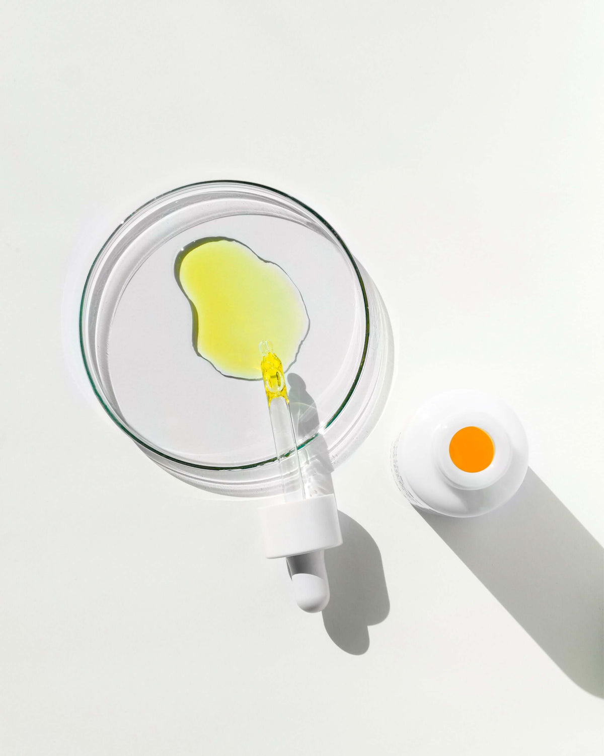 Coupelle en verre avec une flaque d'huile jaune et une pipette, flacon ouvert à côté, vue du dessus