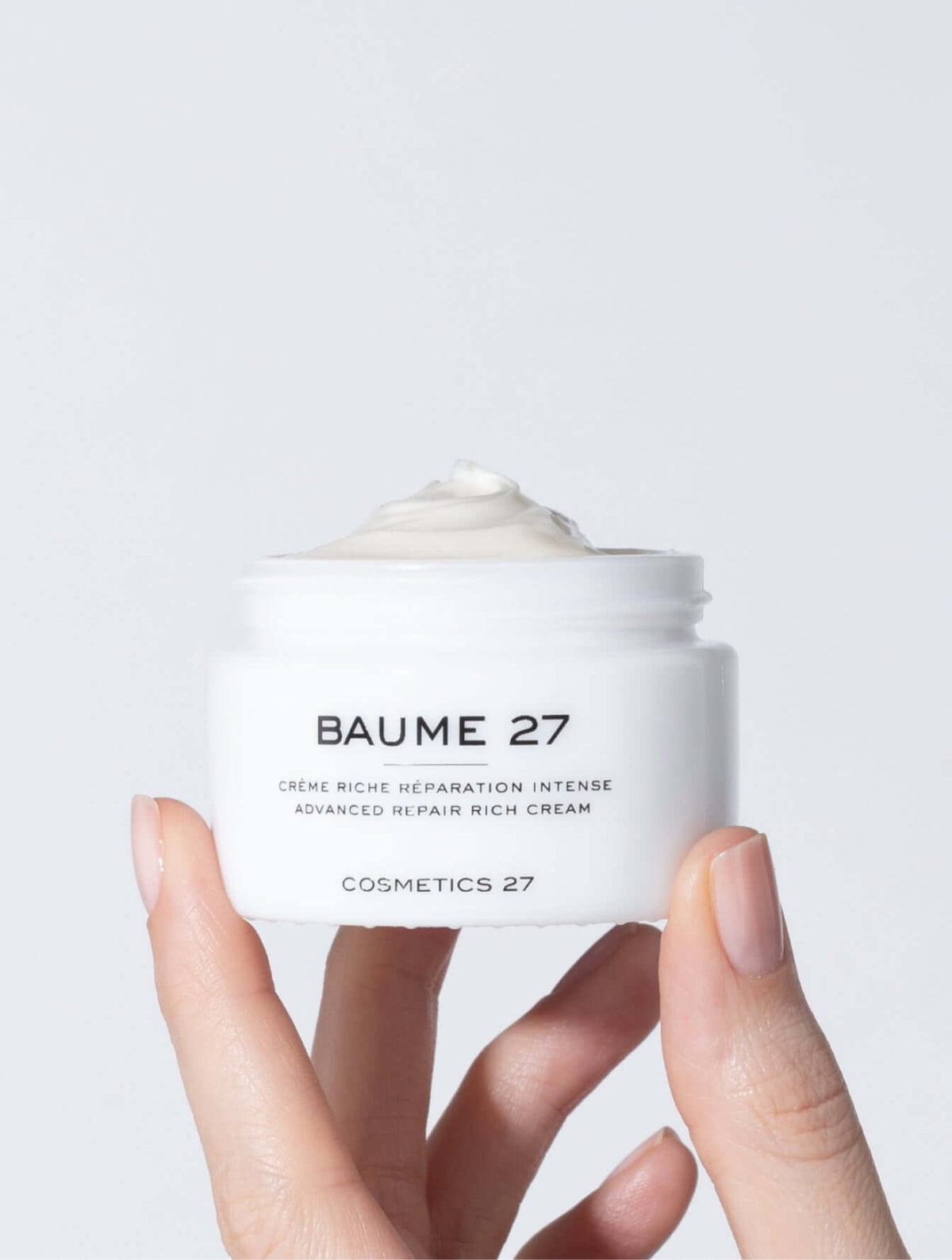Two hands holding an open jar of Baume 27 Rich Cream filled with white cream