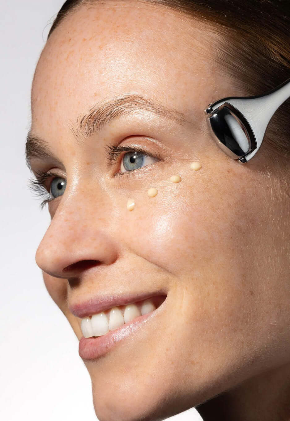 Smiling woman gliding the Ridoki roller near her eye, with dots of cream on her cheekbone