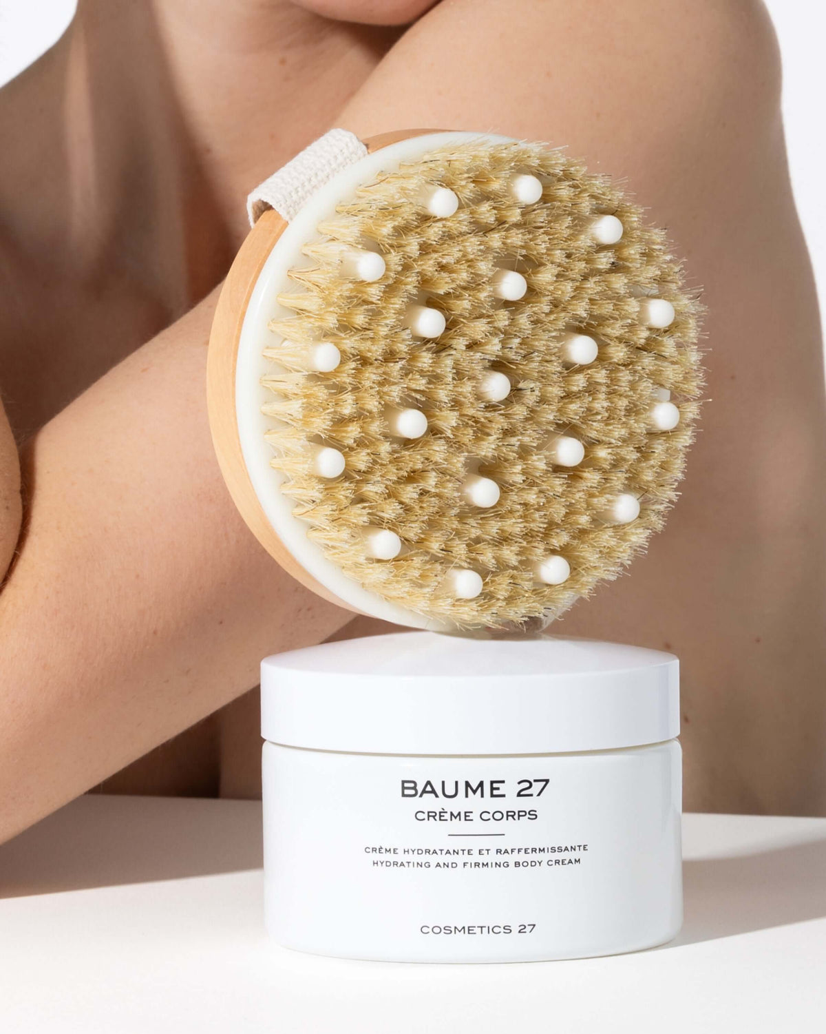Brosse pour le corps tenue contre le bras, pot de Baume 27 Crème Corps au premier plan